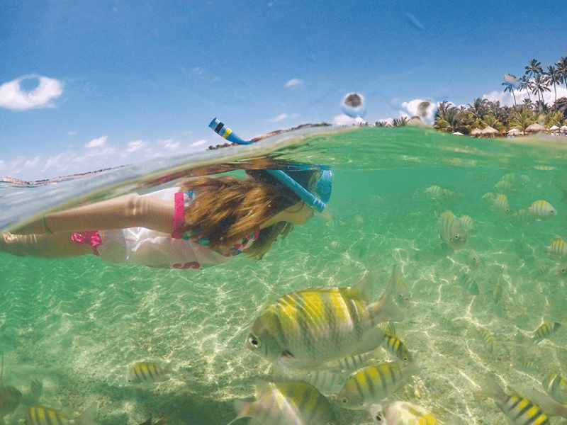 Jovem fazendo snorkeling rodeada de peixes tropicais na cristalina Praia de Serrambi, destacando a rica vida marinha local.