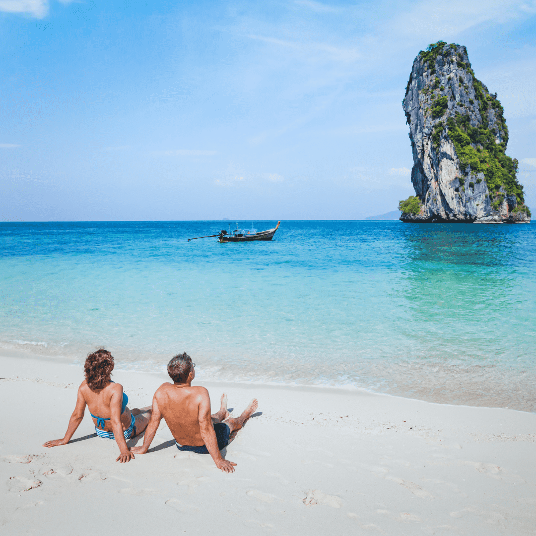 Casal sentado na areia observando o mar azul turquesa e uma ilha rochosa em uma praia tropical