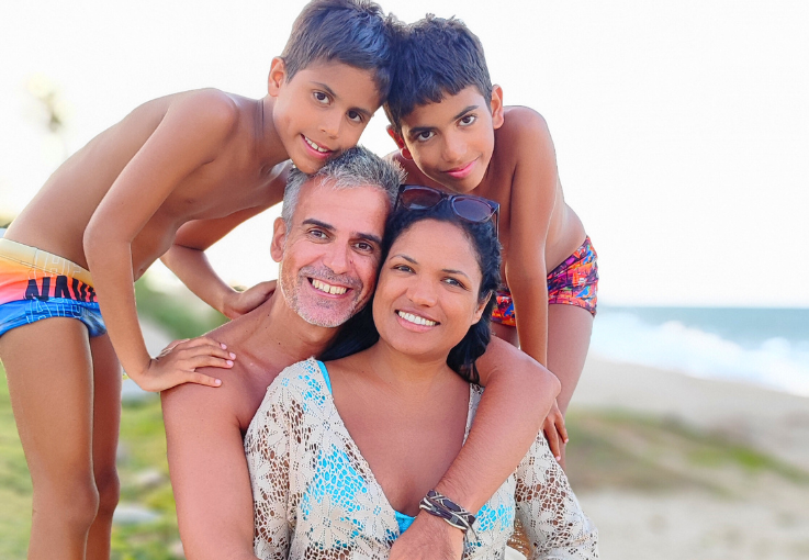 Família Amorim sorridente aproveitando dia ensolarado na praia com mar ao fundo